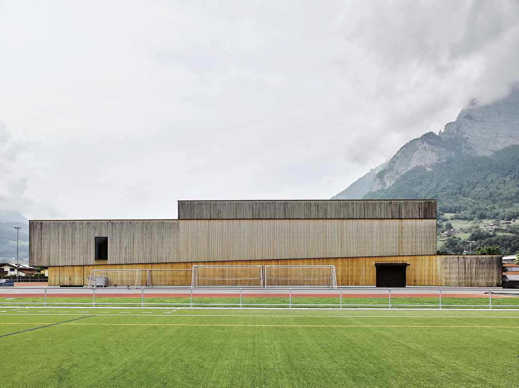 Sporthalle mit Holzfassade an Sportplatz vor Bergen