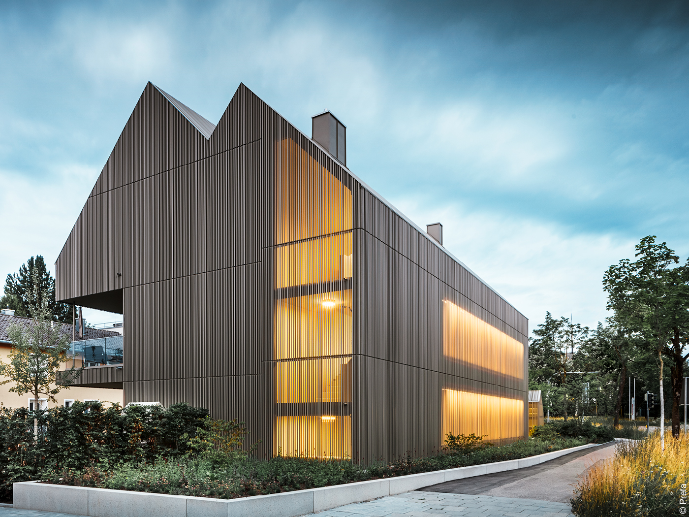 Mehrfamilienhaus mit Aluminiumfassade und heimelig beleuchteten Fenstern vor bewölktem Himmel