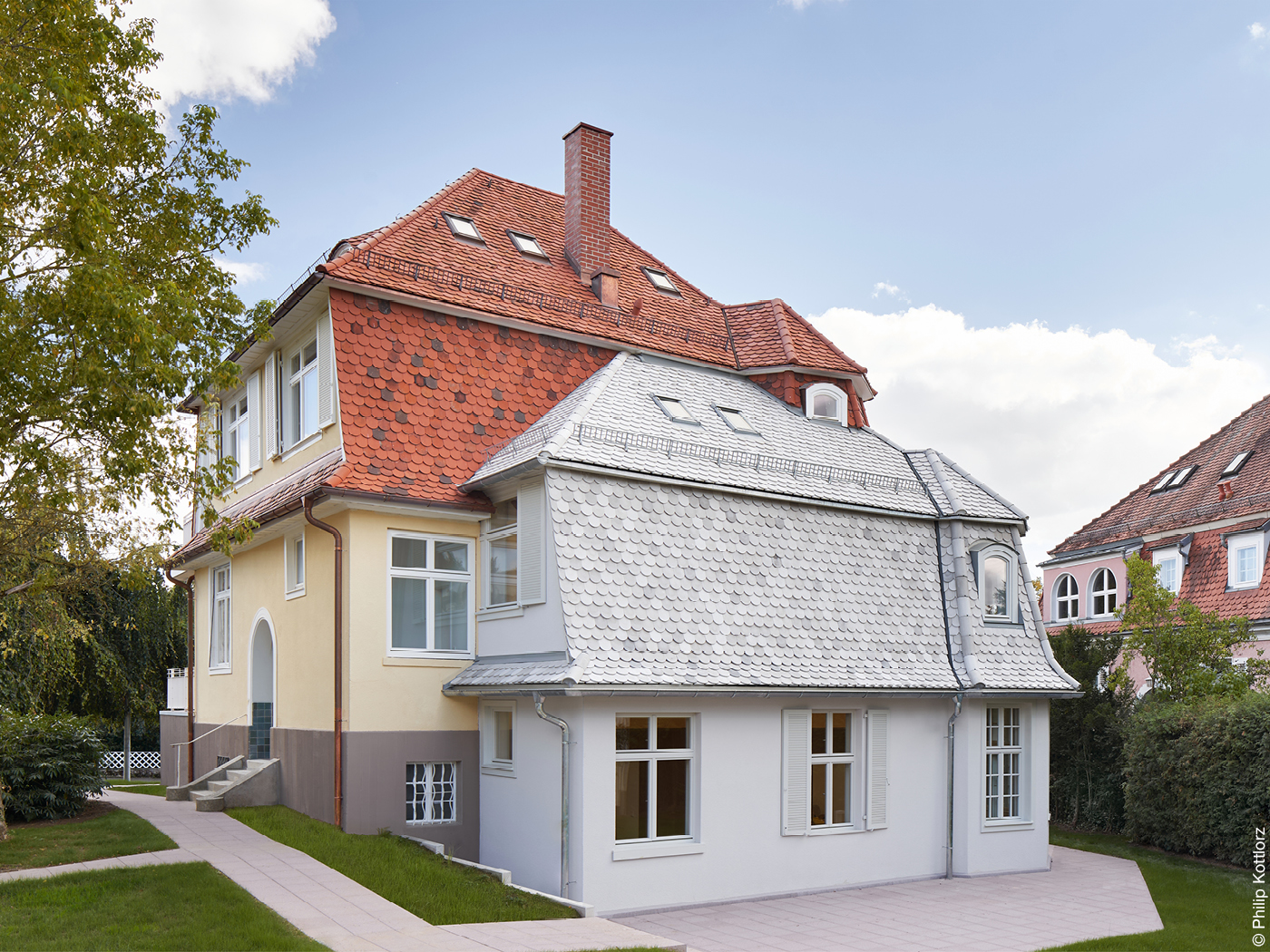 Ein zitronengelbes Haus mit rotem Ziegeldach und einem weißgetünchten Anbau mit weißem Ziegeldach. 