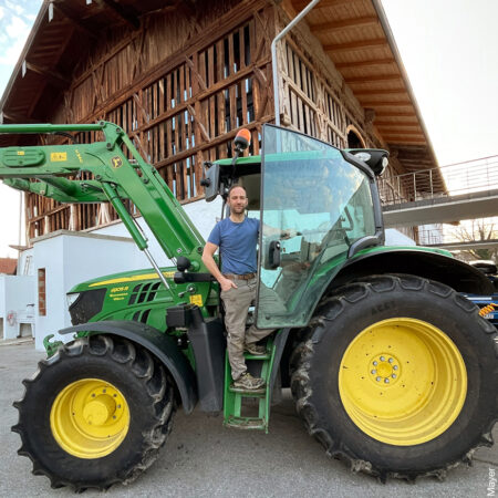 Mann auf einem grünen Traktor vor einem Haus mit Holzfassade.