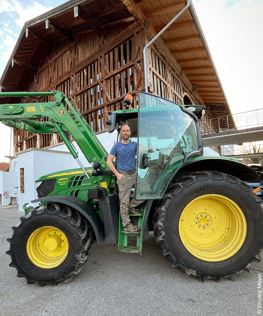 Mann auf einem grünen Traktor vor einem Haus mit Holzfassade.