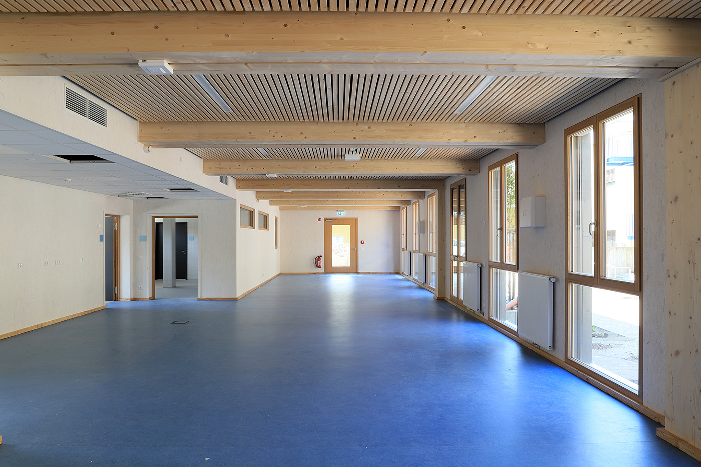 Breiter Schulflur mit blauem Linoleum und bodentiefer Fensterfront.