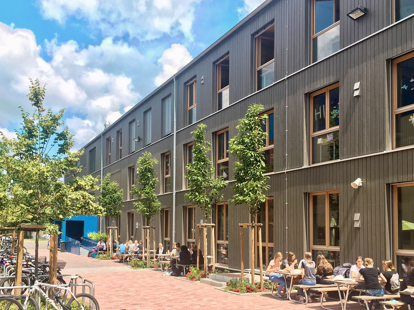 Modulbau-Schule mit Holzfassade, davor sitzen Schüler im Sonnenschein und machen Mittagspause.