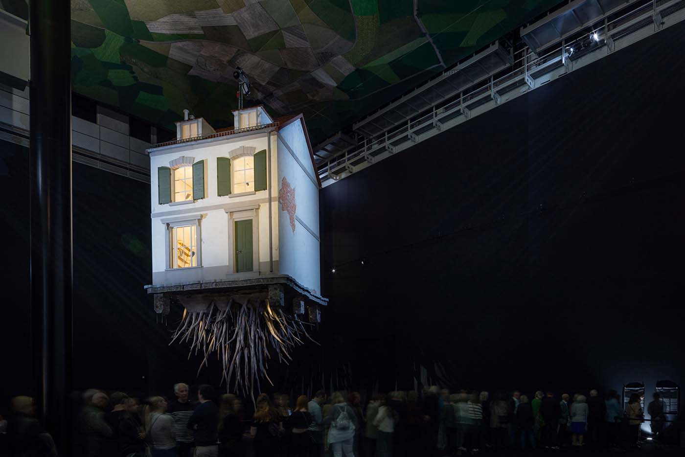 Künstlich installiertes weißes Haus mit Wurzeln hängt an der Decke eines dunklen Innenraums einer Kunsthalle