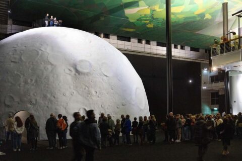 Künstlich installierter Mond in einer dunklen Kunsthalle mit vielen Besuchern