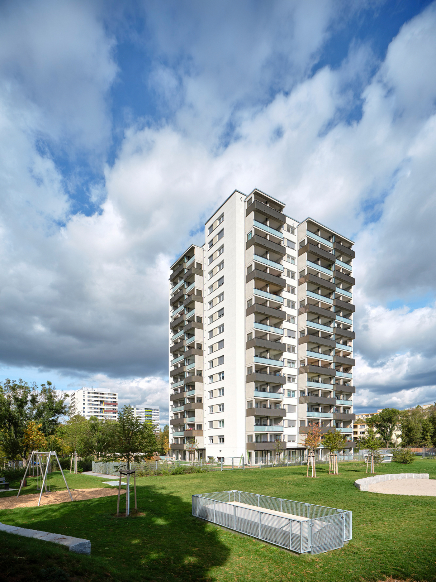 Hochhaus mit weißer Fassade und hellblau-grauen Balkonen auf Rasen mit einem Spielplatz mit Schaukel.