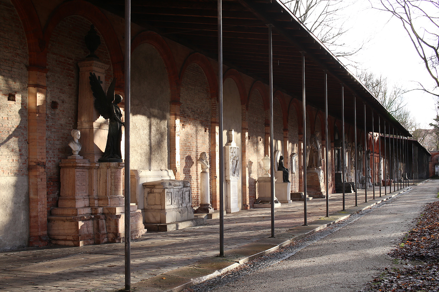 Langer Gang am Münchner Südfriedhof mit Steinsockeln, Büsten und Engelsfiguren.