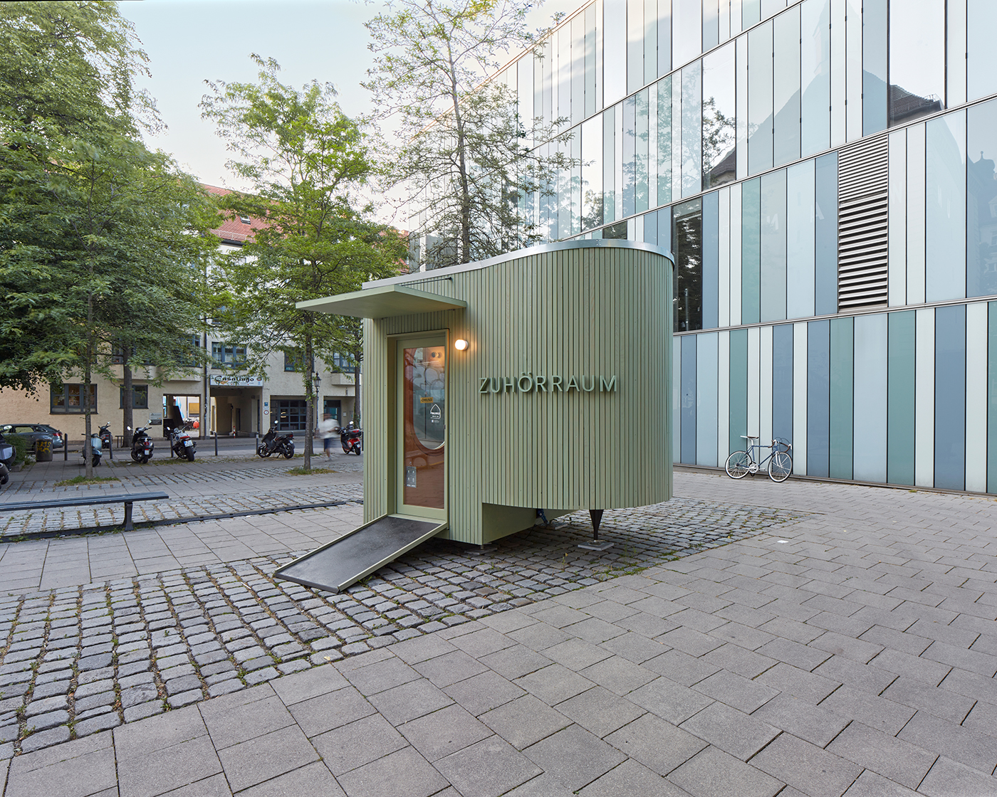 Ovaler Pavillon mit Aufschrift Zuhörraum und lindgrüner Holzverkleidung auf einem städtischen Platz mit Kopfsteinpflaster.